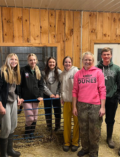 Group of six smiling people, standing inside a barn.