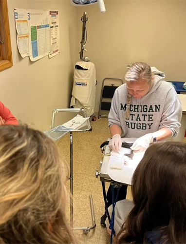 A person in a Michigan State Rugby sweatshirt teaches a small group from a clipboard in a clinical setting.