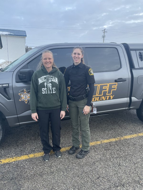 Two women, one in a Michigan State sweatshirt and one in sheriff's uniform, standing beside a patrol van.