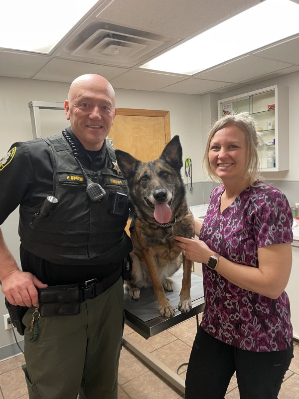 A sheriff and a woman posing with a happy German Shepherd in a vet's office.